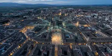 Vista aérea de una ciudad histórica española al anochecer, con techos de teja y plazas, superpuesta con un entramado digital que simboliza la preservación cultural y la tecnología.
